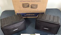 Pair of black Bose 101 Series II speakers with ribbed plastic housing and metal grille showing the Bose logo, positioned in front of the original cardboard box.