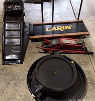 Photo showing black metal creeper with Larin logo next to red hydraulic floor jack, round black oil catcher, ramps, and battery charger