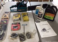 Makita chainsaw with bar and chain, multiple new chains in packaging, Power Sharp diamond sharpener kit, bottles of oil and lubricants, wrench tool, and owner's manual arranged on table.