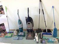 Wide view showing cleaning supplies including steam cleaner, upright floor cleaner, vacuum, brooms, and dust mop.