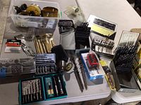 Overview of multiple item groups on a table including drill bit sets, hole saws in plastic container, driver bits and metal holders.