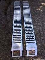 Pair of metal loading ramps laid on asphalt showing perforated design and base with warning labels.