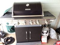Wide view of the grill on wheels with one propane tank, regulator kit box and folded canvas cover visible