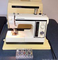Front view of Kenmore sewing machine on beige base with clear plastic accessory box and hard carrying case behind.