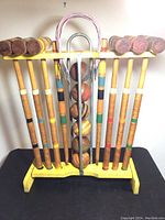 Full view of the vintage croquet set in yellow wooden stand showing six mallets, six balls, two metal wickets, and one wooden stake.
