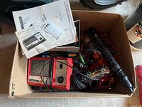 Box containing a red and beige Acrison branded automotive meter with wires, cables, probes and several papers/manuals on top