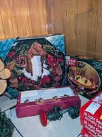 Photo showing Christmas wreath with red ribbons and ornaments, Mr. Christmas Christmas Eve Ball in box, partially coiled string lights with green wire and red bulb covers, and a box of stocking hangers