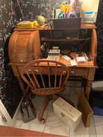Front view of wooden roll-top desk and matching wooden office chair surrounded by office supplies. Desk includes roll-top cover, small drawers, and pigeonholes.