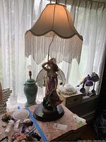Full view of ceramic figural lamp with figurine of woman in pink dress under beige lampshade with fringe.