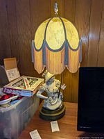 Full view of the vintage figural lamp with lampshade, decorative fringed edge, illuminated indoors on wood table.