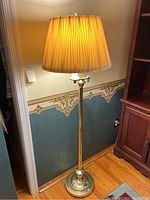 Full view of the floor lamp featuring the brass tone pole and pleated fabric shade, lit and standing on a hardwood floor near a wall with patterned wallpaper.