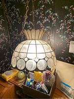 Photo showing the front view of the vintage swag hanging lamp illuminated, highlighting the translucent capiz shell panels and gold-tone metal frame with scalloped design at the top and round panels around the center.