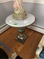 Front angle showing round white marble top with brass edge and ornate brass pedestal base with detailed floral designs, sitting on wood floor near wall baseboard.