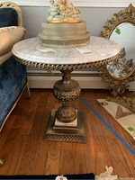 Front angle showing round white marble top and ornate metal base with floral and scrollwork detailing.
