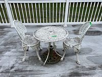 Full front view of white cast metal patio bistro table with two matching chairs and umbrella stand on weathered outdoor deck.