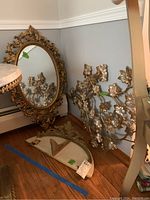 Photo showing the ornate oval mirror with gold carved frame, metal floral wall decor with flowers and leaves, and decorative shelf on a wood floor.