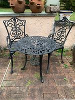 Metal patio set with one round table and two chairs outdoors on a brick surface, showing ornate scrollwork design.