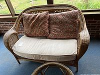 Full front view of the rattan loveseat with beige seat cushion and two patterned accent cushions on a covered patio.