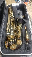 Jean Baptiste JB-480A black alto saxophone body inside open hard shell case with black plush interior, gold plated keys visible alongside neck and mouthpiece.
