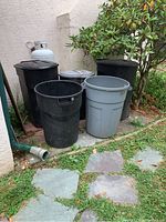 Outdoor photo showing five plastic trash bins placed on a stone slab near a wall and a small tree. Bins vary in size, color, and style; some have lids and others do not.