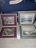 Four framed pieces of wall art including two textile needlework scenes and two watercolor paintings, displayed leaning against a couch