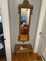 Full length view of the ornate gold framed mirror hung on the wall above the padded bench seat with gold finish and cabriole legs