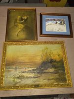 Photo showing three framed artworks: the top left is 'HOPE' print with a seated figure in gold frame, top right is a small snowy winter rural scene, the bottom artwork is a large seascape oil on board with boats and rocky shore in a thick gold frame.