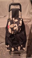 Photo of the front view of the black and beige floral patterned shopping cart with handle and flap closed.
