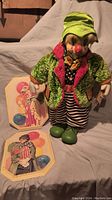 Front view of 20-inch clown doll in colorful outfit next to two wooden framed clown artwork pieces on fabric background.