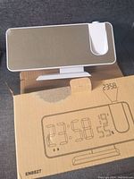 Front view of the digital projection alarm clock placed on top of its box. Mirrors front display with protective plastic still attached.