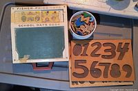 Top view of vintage Fisher-Price School Days Desk showing chalkboard surface with signs of wear and colorful school-themed illustrations on frame. Number cutouts and storage container beside the desk.