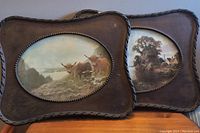 Two old wooden oval frames with pictures inside, one featuring cows in a pasture and the other a riverside scene.