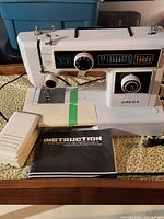 Off-white Omega sewing machine with stitch pattern selection and control dials, foot pedal and manual visible on wooden table.