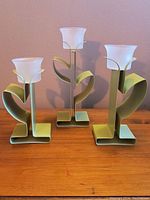 Three green metal candle holders with frosted glass flower shaped cups placed on wooden surface against light colored wall