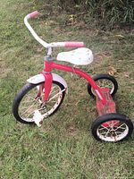 Side angle view of red and white metal tricycle showing front wheel, white seat, red frame and wear on paint and rust on metal parts.