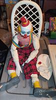 Photo of sitting clown doll showing seated position on white lattice chair with visible damage to hat and hand.