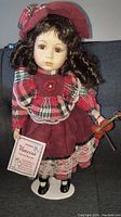 Full view of Vanessa porcelain doll wearing burgundy and plaid dress, holding violin, standing on white stand with certificate tag.