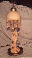 Front view of vintage Crosa Collection lady holding peacock table lamp with beaded fringe shade on wooden base