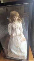 Full view of the porcelain doll in a clear plexiglass case, showing the detailed white dress and facial features.