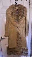 Front view of vintage long leather jacket with thick brown fur collar and fur trim along hem, hanging on a hanger against a white door.