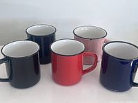 Front-left angled view of the five ceramic mugs showcasing the enamel look and rust detail at rims, arranged in a staggered formation.