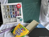 Green cutting mat with grid, Crayola colored pencils box, knitting needles, canvases and wood panels, and the glitter mixing kit in its packaging.