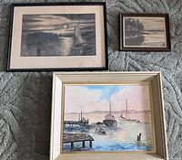 Three framed nautical themed artworks displayed on carpet: black and white sailboat sketch, small black and white landscape, and watercolor boats by pier.