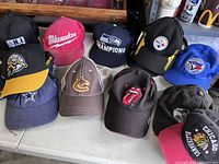 Photo showing eleven assorted ball caps placed on a table, featuring logos from various sports teams and band.