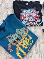 Both folded T-shirts, Party Hat shirt in blue front visible, Tilt shirt underneath folded with graphic visible.