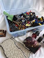 Plastic bin filled with assorted Lego bricks and accessories, loose in various colors with black roof tiles, green foliage pieces, and dinosaur figures visible