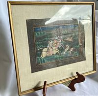 Framed hand painted silk artwork depicting a hunting scene, viewed angled on wooden display stand, showing gold frame and beige mat.