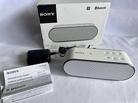 White Sony SRS-X2 speaker alongside power charger, user manual, and original product box