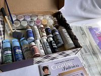 Overview of the stained glass art making kit contents inside open box, showing multiple bottles of different color paints, brushes, and a book titled 'Creative Interiors'.