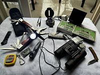 Overall view of survival gear items laid out on white cloth, showing tools, binoculars, matches, compass, flashlight, padlock, and other accessories.